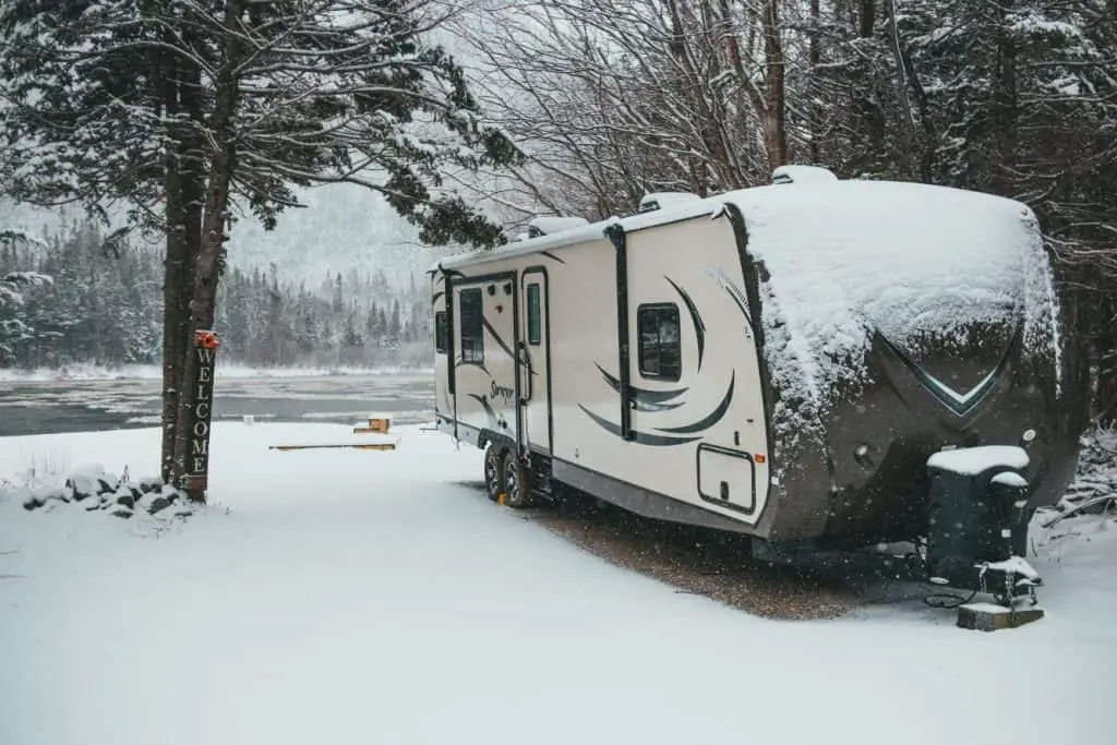 DIY RV skirting ideas for winter camping and full time RV living: image shows a front shot of a travel trailer with snow on the ground and on the roof. in the background is a stream and snow covered forest. there is a sign leaned up against a tree in the foreground that says "welcome"