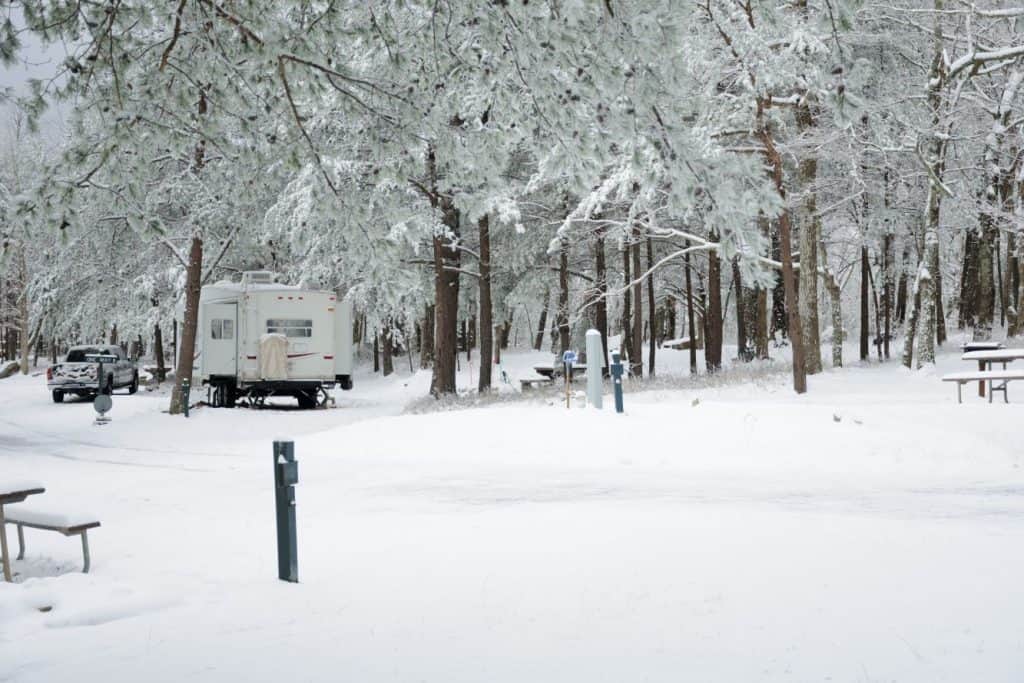 living in fifth wheel in winter: image of a fifth wheel camper parked at a campground with the ground, trees, and picnic tables in the scene all covered in 4 inches of snow